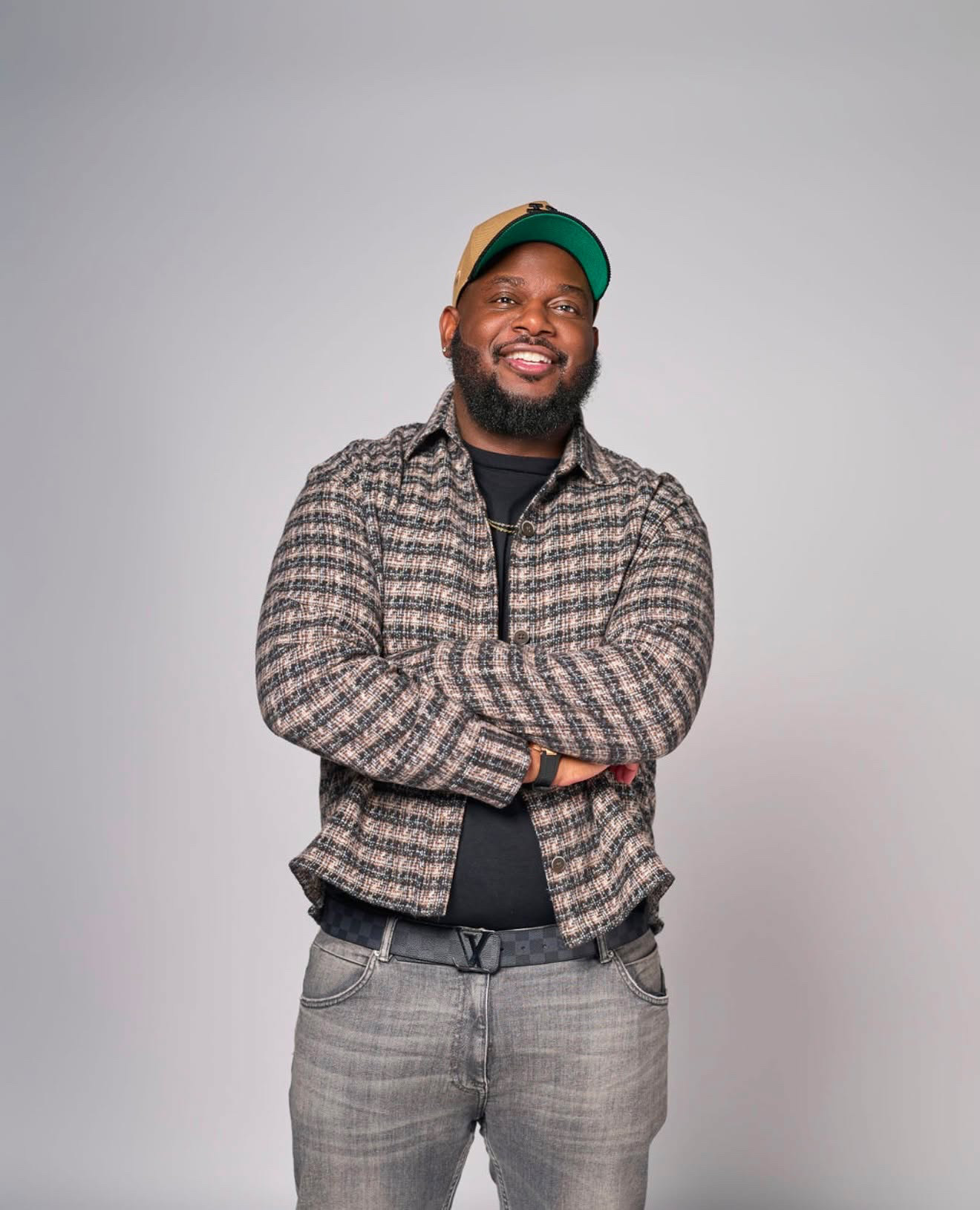 Comedian BTIDY (Brian Tidwell) standing with his arms crossed, smiling, in a checked overshirt and green-and-tan cap.